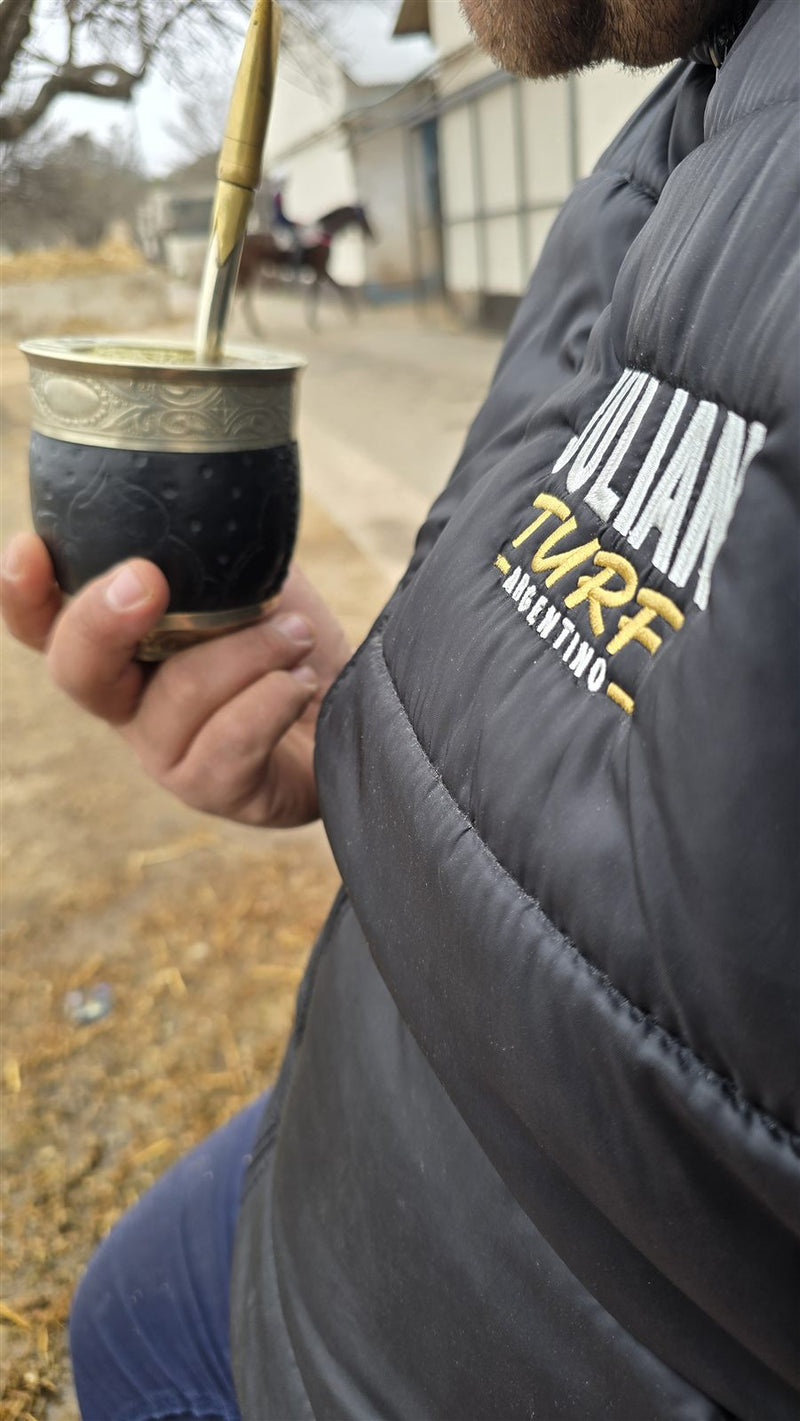 Julián tomando mate en el stud con la campera de Julian Turf mientras pasa un caballo de carrera